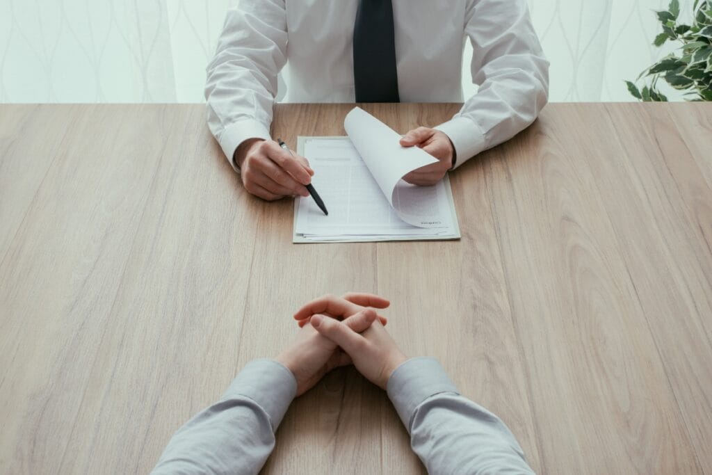 Two people seated at a wooden table, one handing over a document and pen while the other's hands are clasped together, perhaps signaling the importance of comprehensive background checks in sealing the deal.