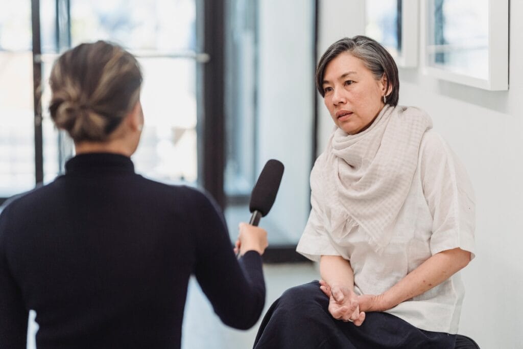 A person holding a microphone conducts an interview with another sitting with arms crossed, wearing a white blouse and scarf. They discuss the importance of comprehensive background checks, highlighting their role in ensuring security and trust indoors.