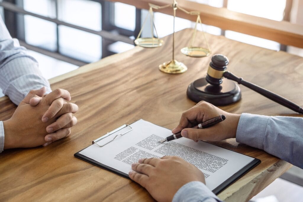 Two people sit at a table, one pointing at a document on a clipboard, discussing comprehensive background checks. In the background, a gavel and scales of justice subtly underscore the gravity of their conversation.