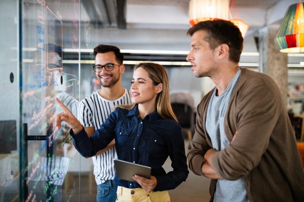 Three people discussing and pointing at data on a transparent board in a modern office setting, emphasizing the importance of comprehensive background checks. One holds a tablet.