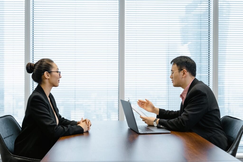 Two people in business attire engage in a discussion across a table, bathed in soft light filtering through the blinds of an office with large windows. The conversation focuses on comprehensive background checks to ensure trust and reliability.