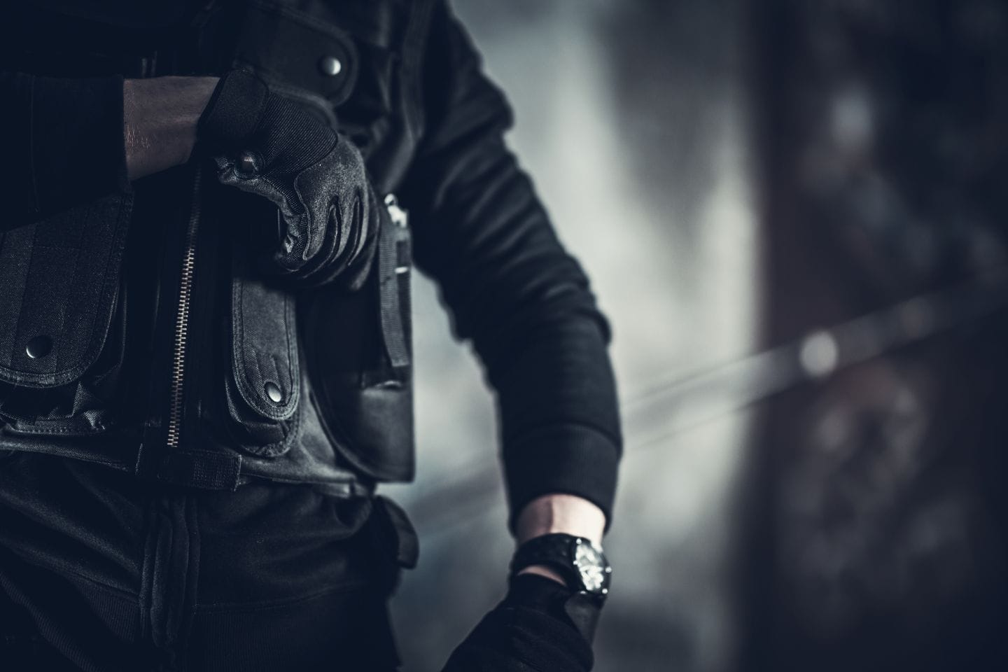 Person in dark tactical clothing, wearing gloves and a watch, holding a small rectangular device or object against a blurred background.