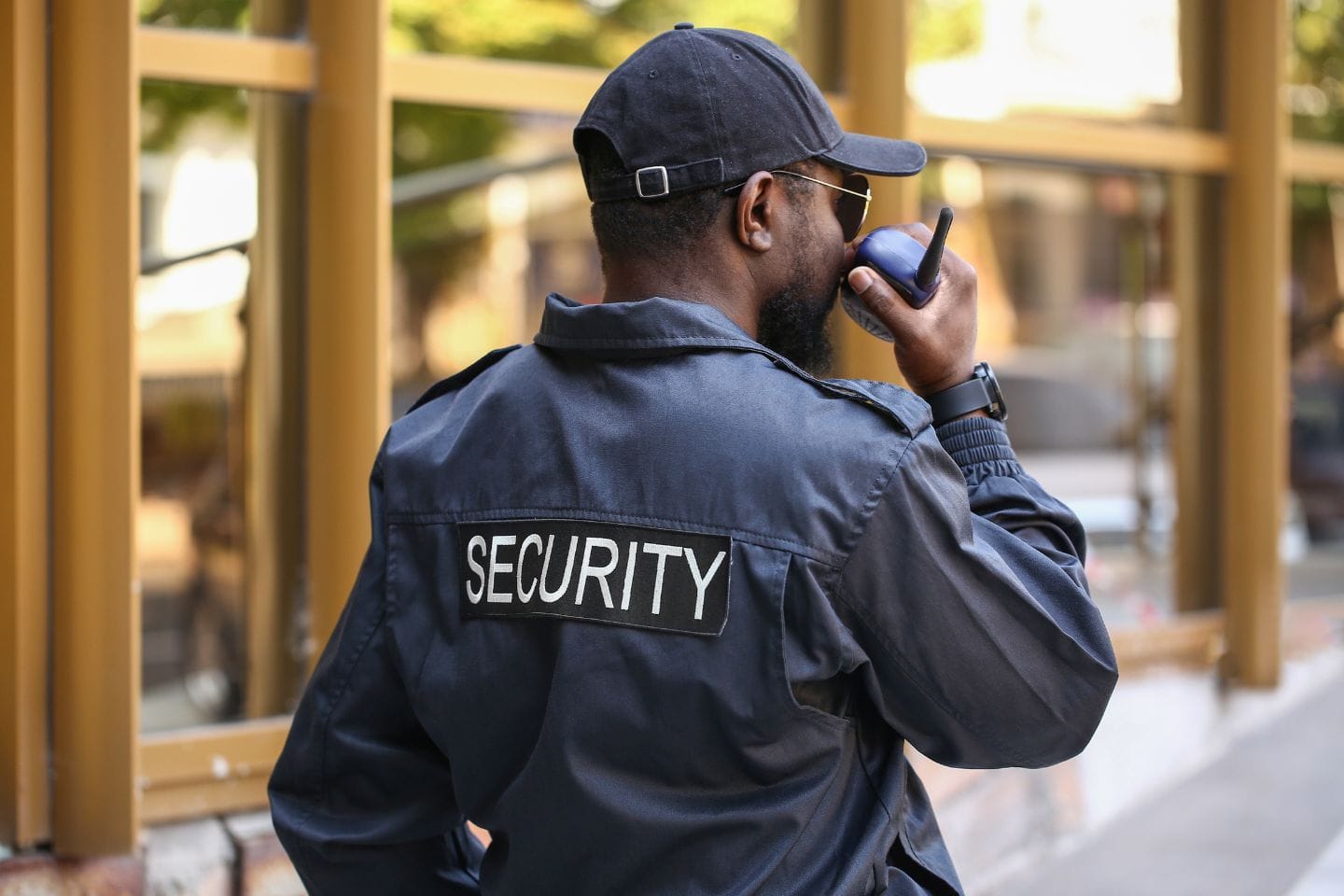 A security guard wearing a cap and sunglasses speaks into a handheld radio while standing outside in front of a building.