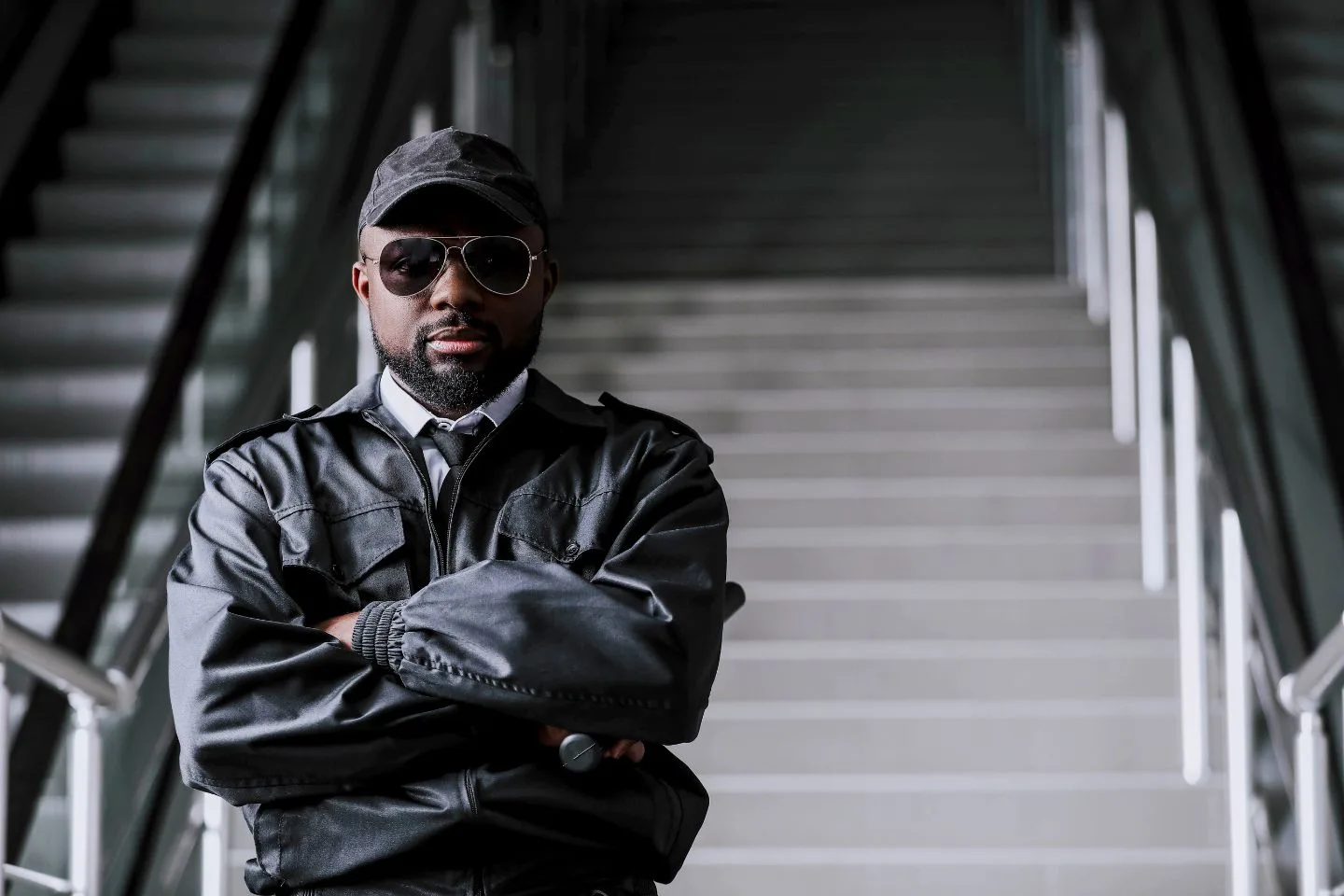 A man in a leather jacket and sunglasses stands with arms crossed in front of a staircase.