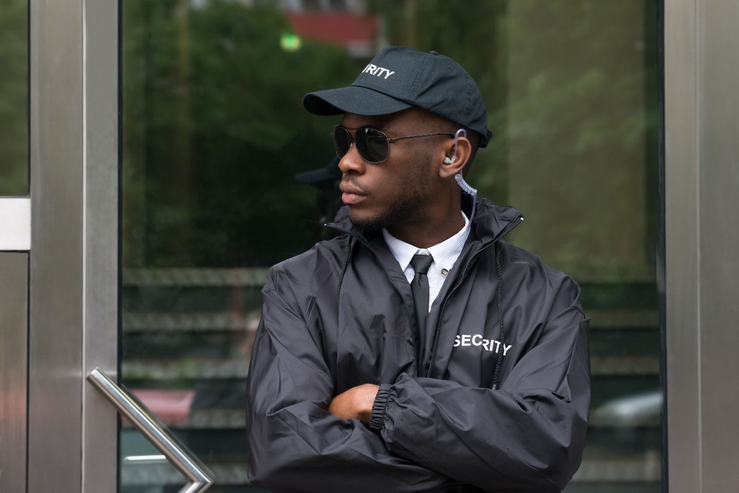 Security guard wearing a black jacket, cap, and sunglasses stands with arms crossed in front of a glass door.