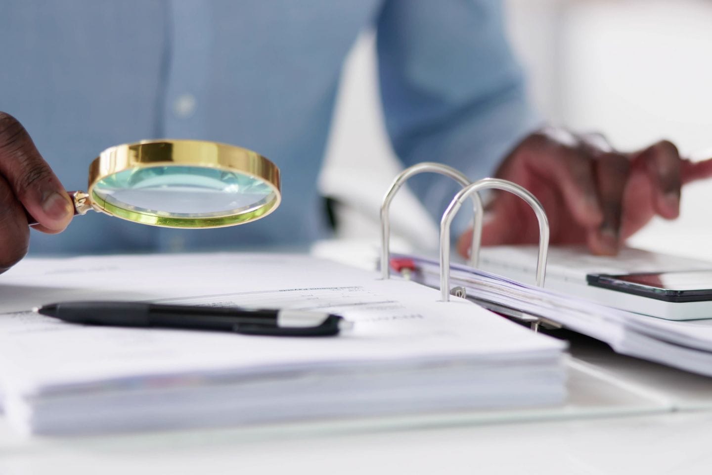 Person examines documents with a magnifying glass, alongside a pen and a smartphone, in a binder.