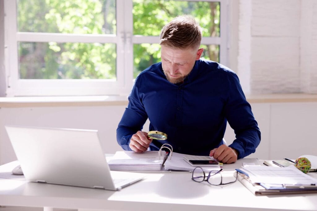 A man in a blue shirt examines documents with a magnifying glass at a desk with a laptop, glasses, and papers. Large windows show a view of greenery outside.