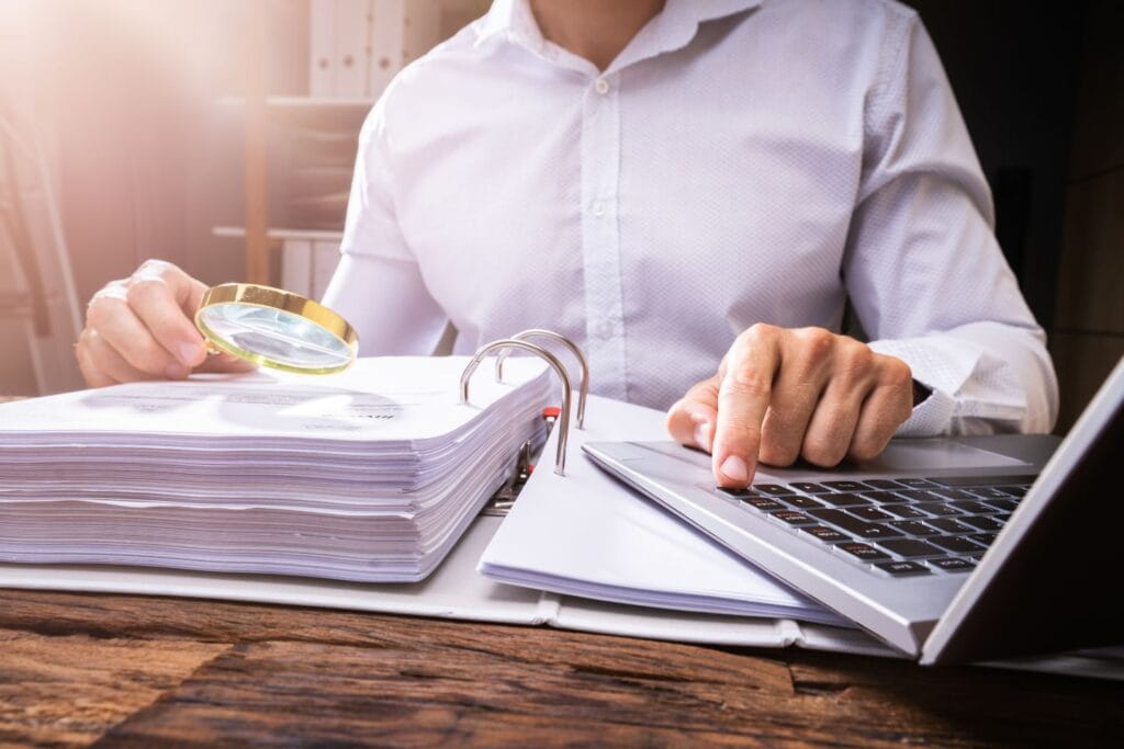 Person examining documents with a magnifying glass, using a laptop, at a wooden desk.