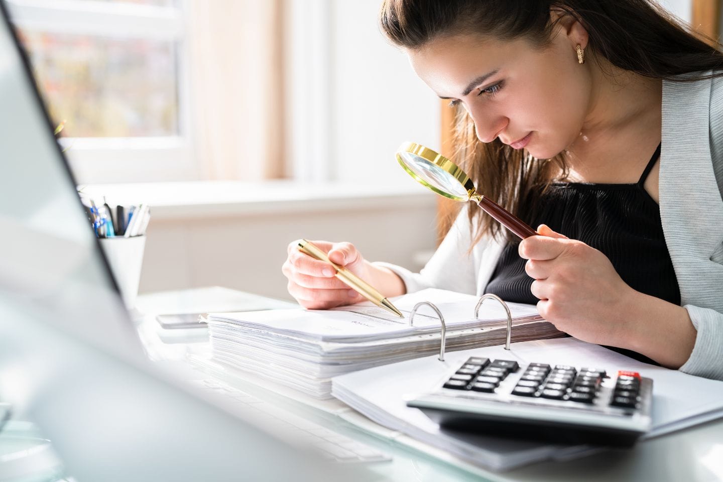 A woman closely examines paperwork with a magnifying glass at a desk, surrounded by binders, a pen, and a calculator.