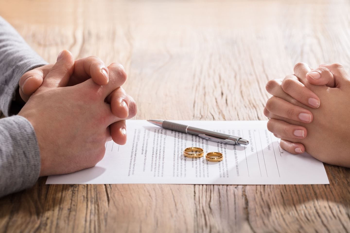 Two people sit across a table with folded hands. Between them are a document, a pen, and two gold wedding rings.