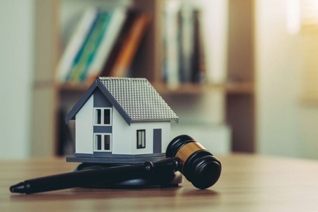 A miniature house sits on a wooden table beside a gavel, with a blurred bookshelf in the background.