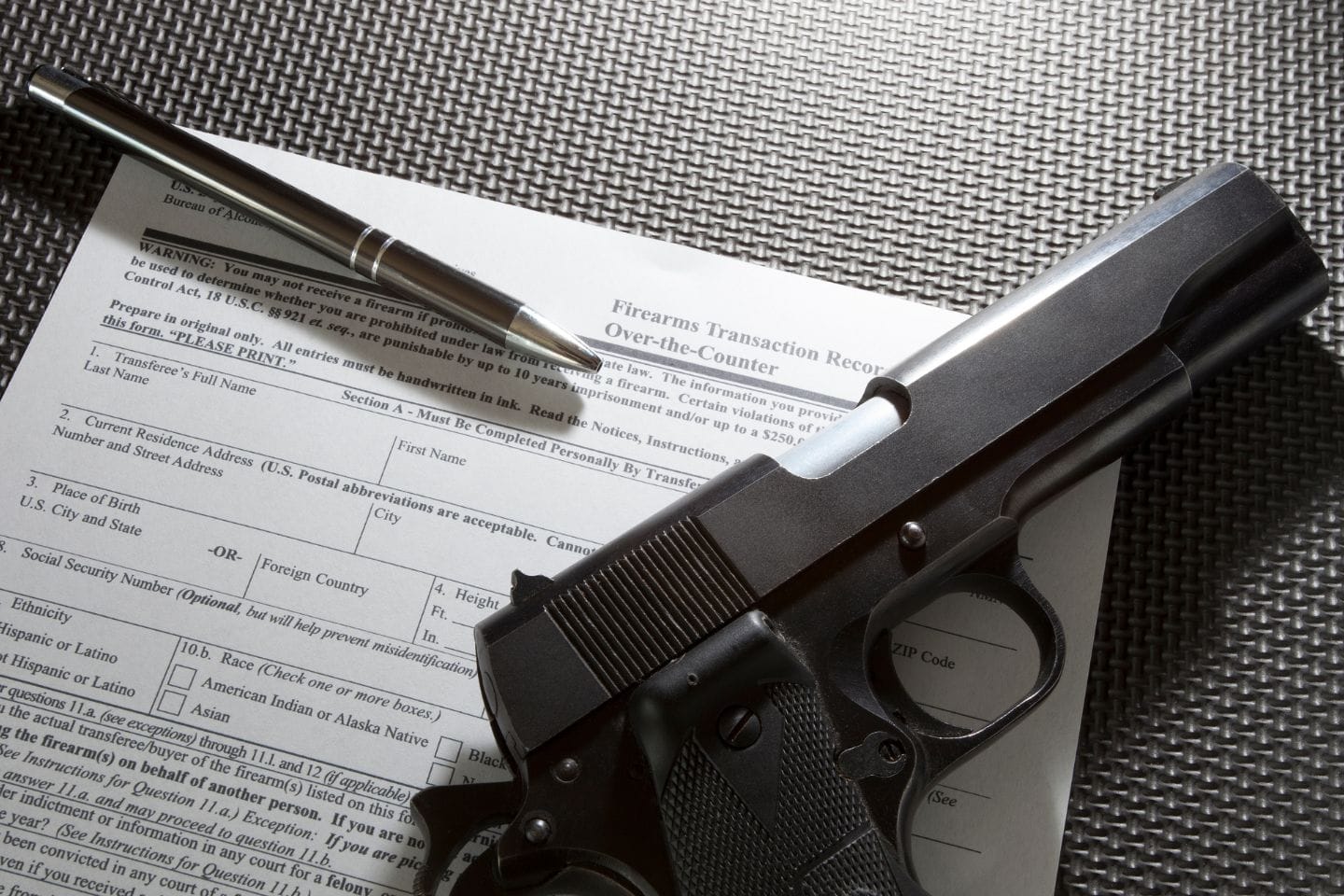 A handgun and pen rest on a firearms transaction record form placed on a textured surface.