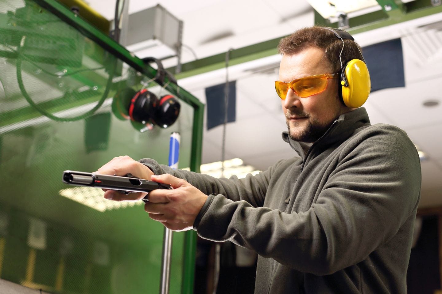 A person wearing ear protection and safety glasses is handling a handgun at an indoor shooting range.