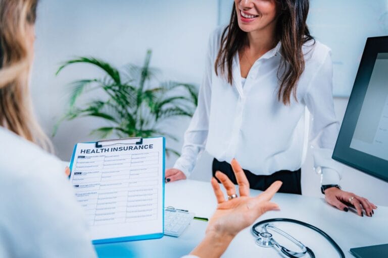 Two people discuss health insurance in an office, exploring various industries served. One holds a clipboard with a form, while a stethoscope and computer are visible on the desk.