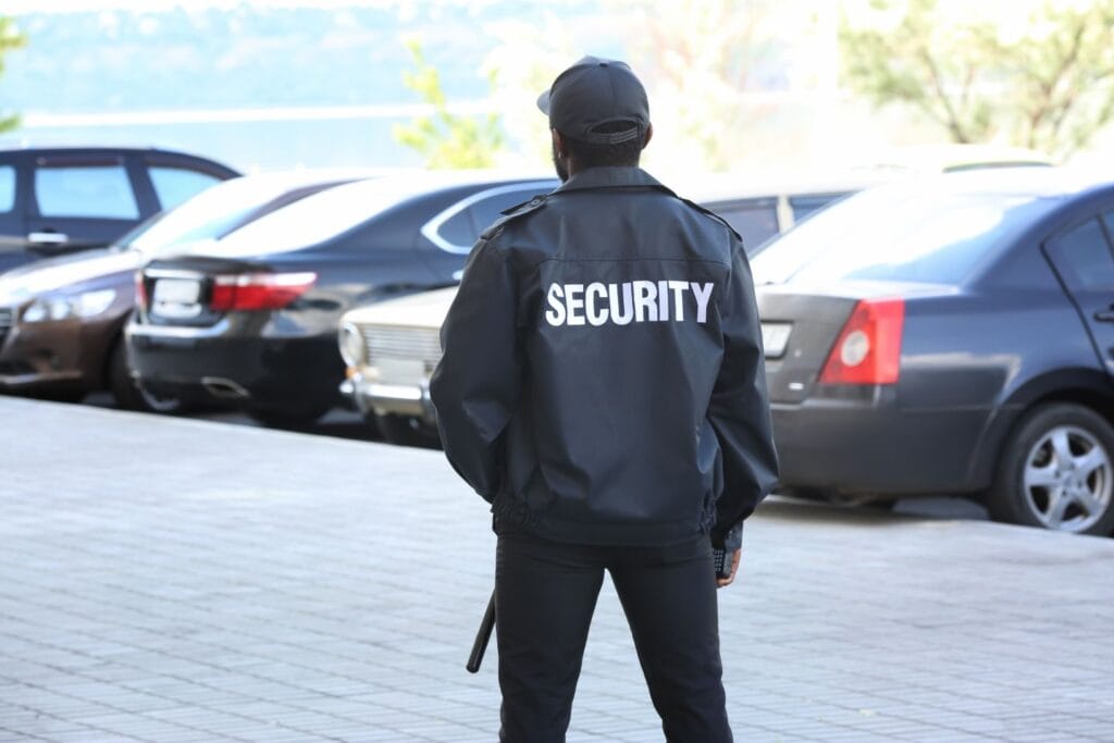 A person in a black security jacket stands in a parking lot, facing away, with several parked cars in the background.