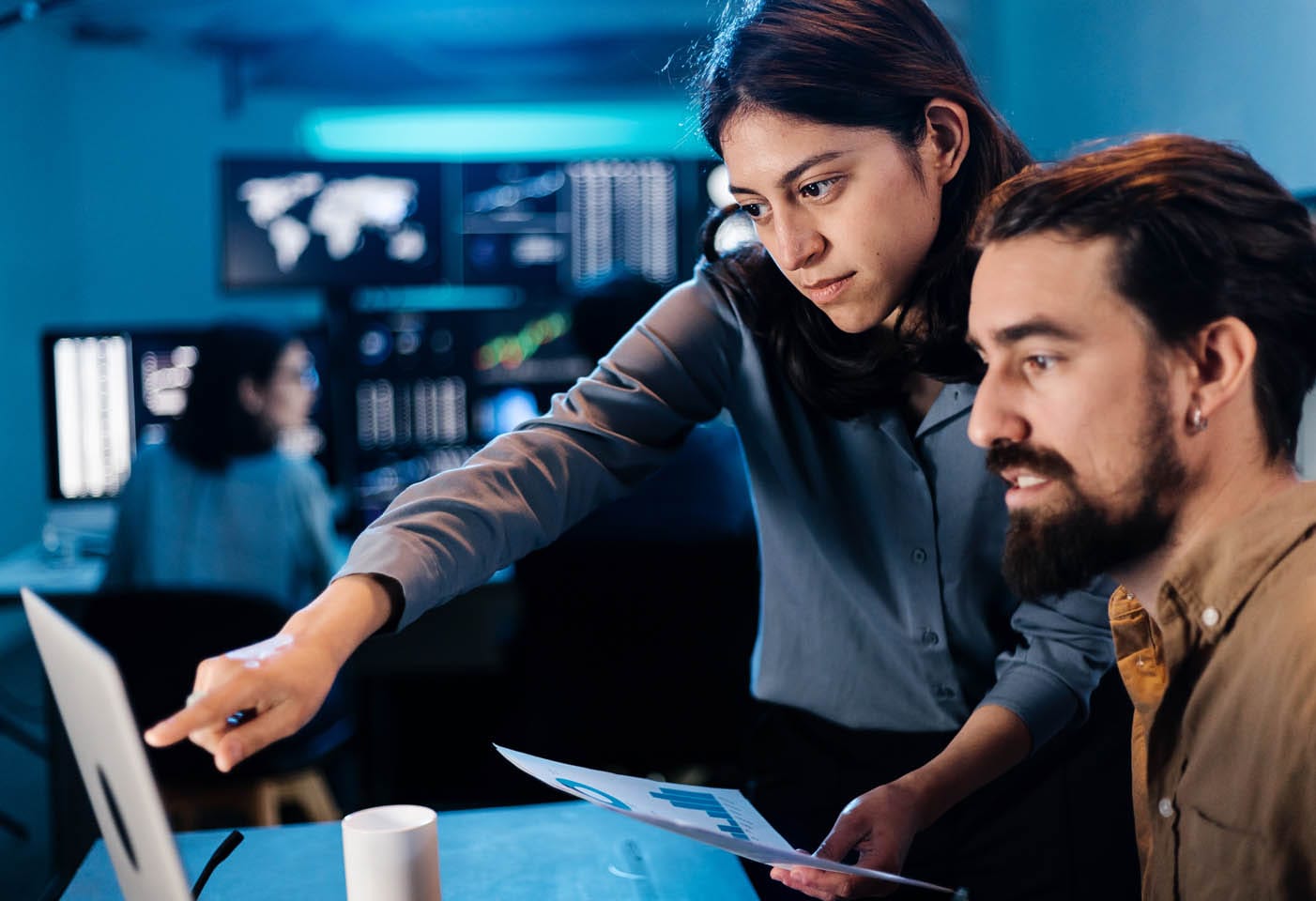 In a bustling office, two people engage in security assessments as they examine data on a computer screen. A woman points intently at the screen while her colleague looks on. Several monitors and colleagues are visible in the background, adding to the atmosphere of focused analysis.