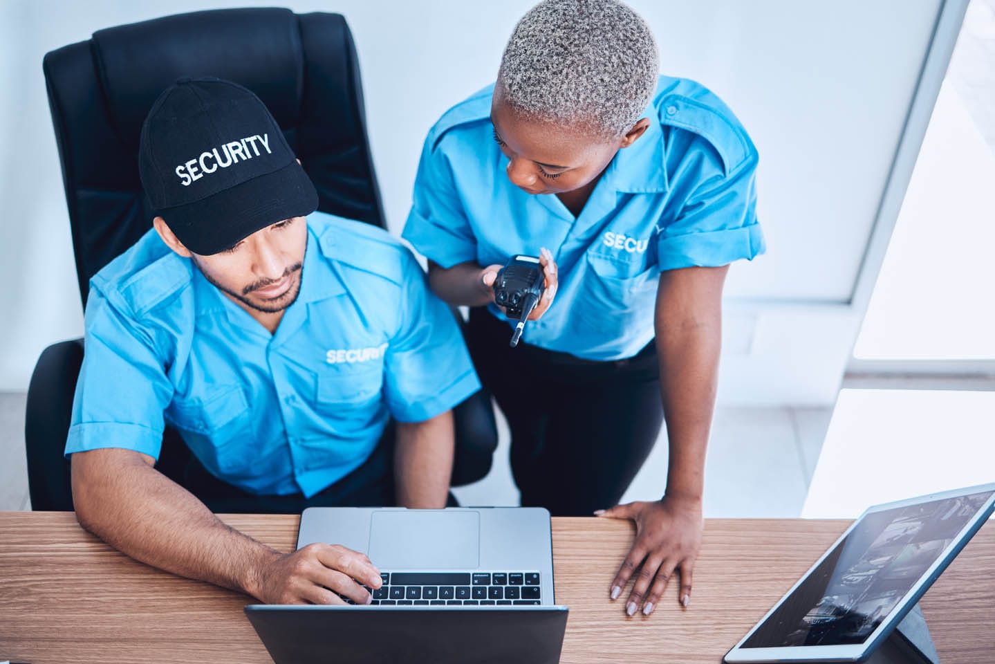 Two security officers in blue uniforms work at a desk with a laptop and a tablet. One is seated, typing out threat assessments, while the other stands holding a radio.