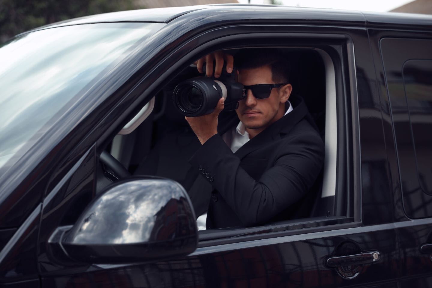 A man in a black suit and sunglasses sits inside a vehicle, holding a camera with a large lens, looking out of the window.