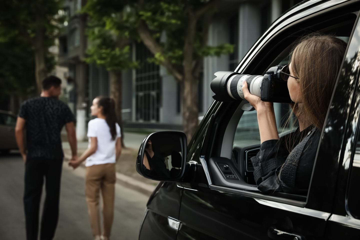 Person in a car uses a camera with a long lens to photograph a couple walking hand in hand on a street.