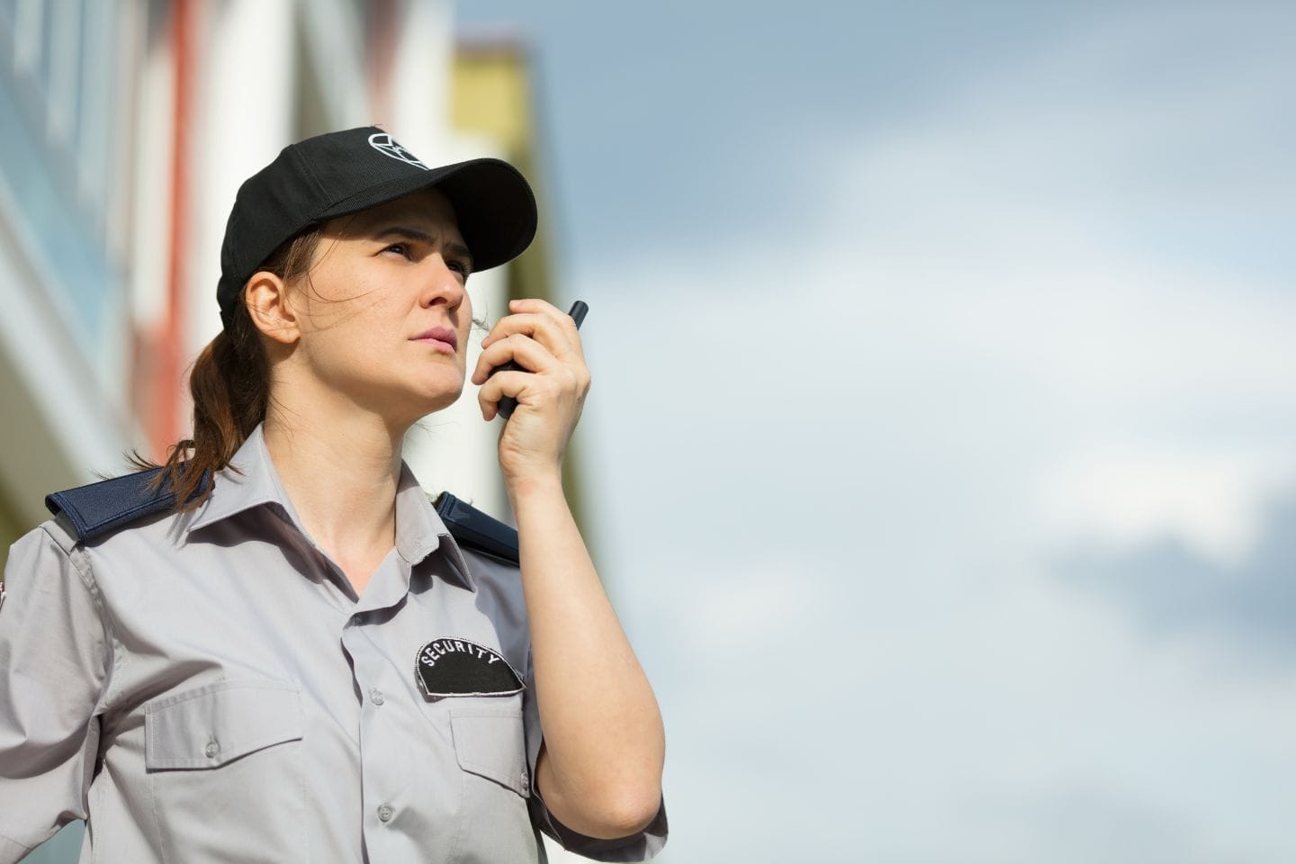 A security guard in uniform, wearing a cap, communicates through a handheld radio while standing outdoors.