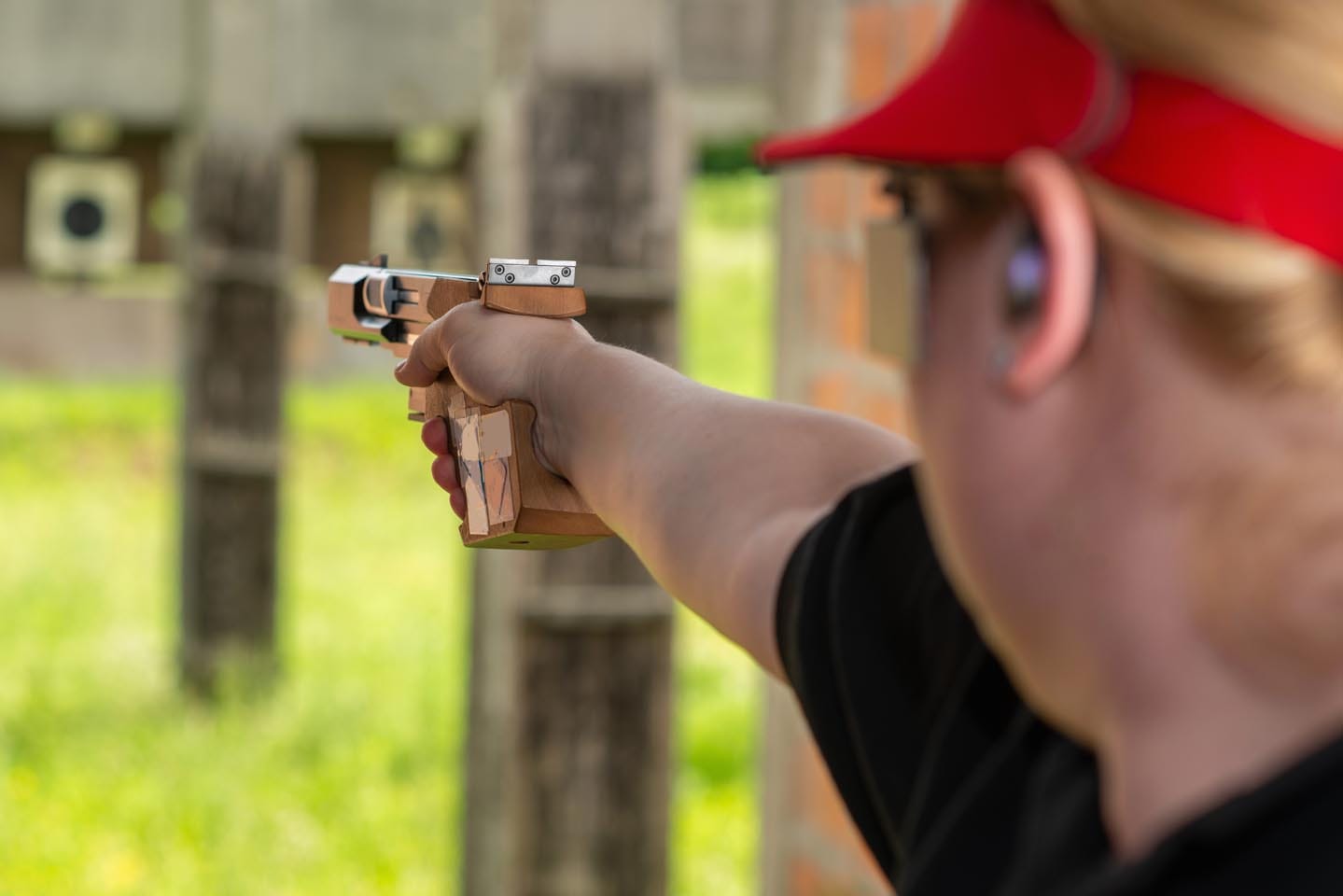 Wearing a red cap and ear protection, the person focuses intently while aiming a pistol as part of active shooter response training, with targets lined up in the background.