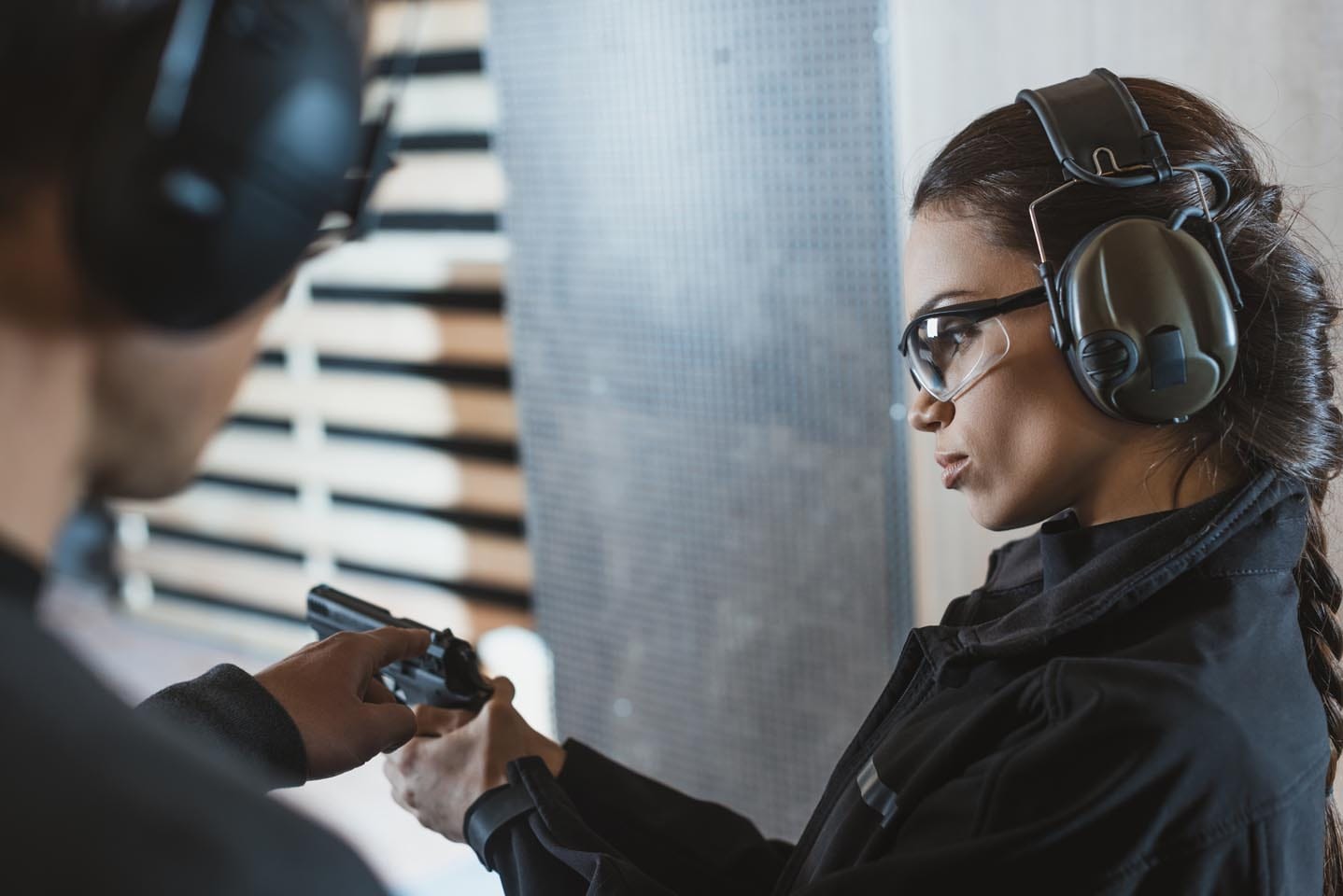 At a shooting range, one person is holding a firearm while the other, equipped with protective glasses and earmuffs, monitors attentively as part of an active shooter response training.