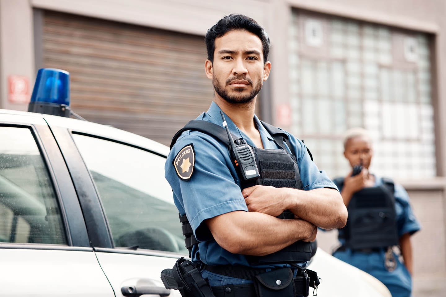 A police officer stands with arms crossed in front of a patrol car, embodying the vigilance typical of armed security. Another officer is seen in the background speaking into a radio, coordination evident in their efficient communication.