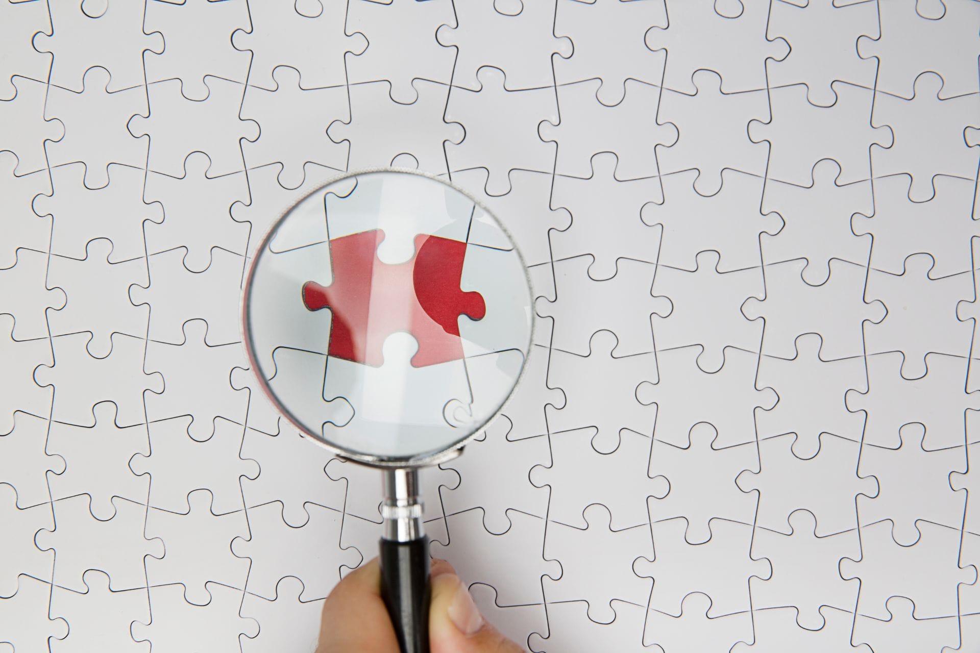 A hand holds a magnifying glass over a white jigsaw puzzle, zooming in on a single red piece.