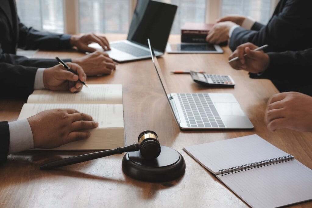 Four people at a conference table with laptops, books, a gavel, and a notepad, appear to be in a meeting or legal discussion.