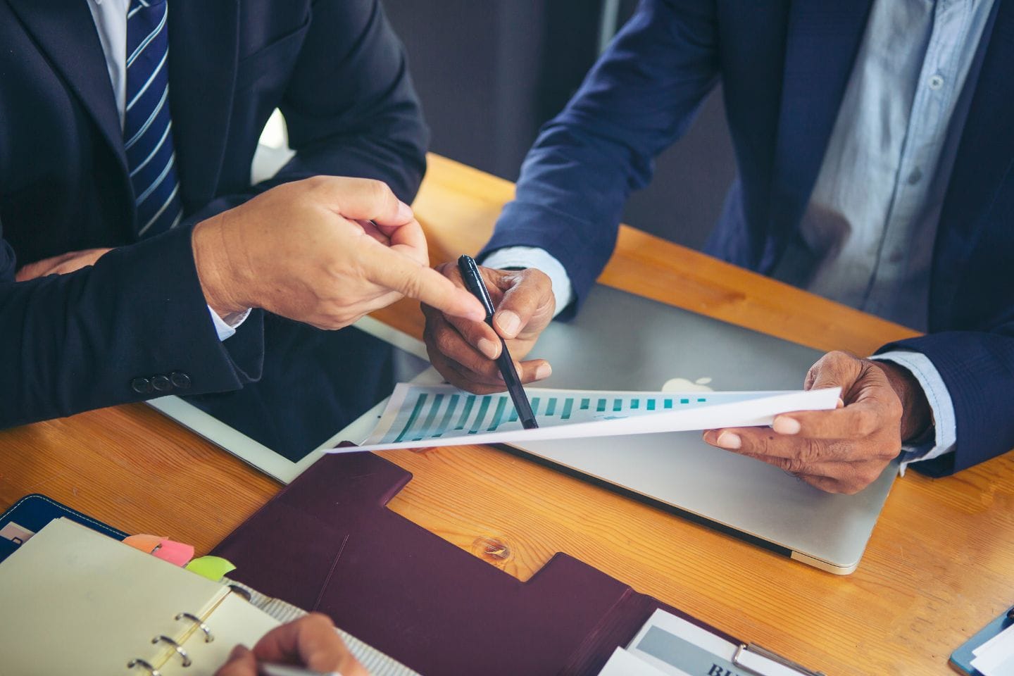 Two corporate professionals in suits are engaged in a detailed discussion about a bar graph at a wooden table, surrounded by a laptop and documents.