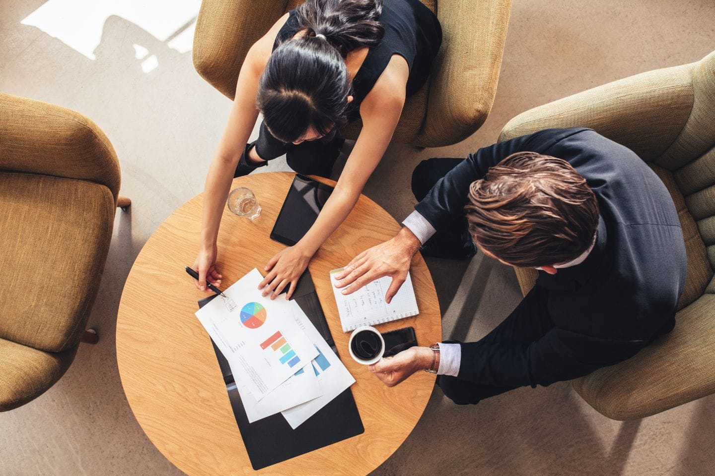 Two people sit at a round table, engrossed in reviewing charts and graphs. One holds a pen, the other cradles a coffee cup, as they delve into the details of corporate investigations.