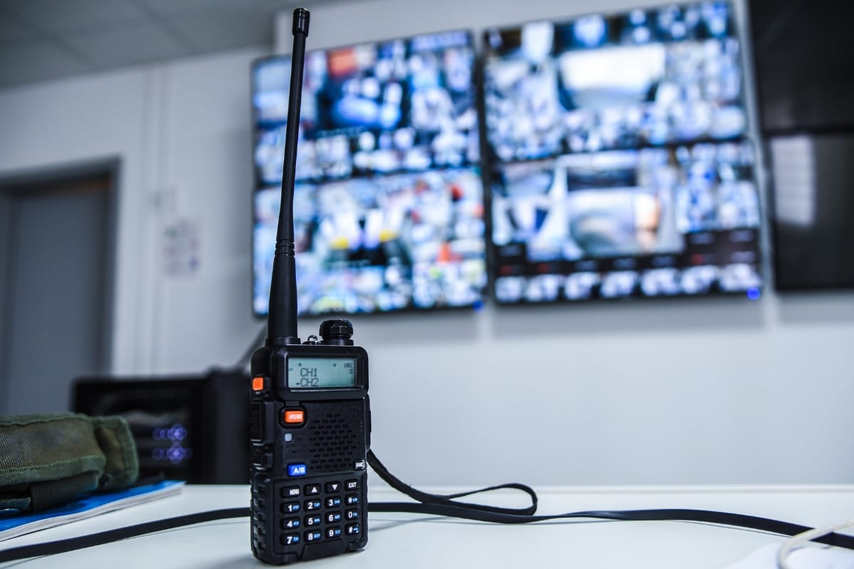 A walkie-talkie rests on a table, crucial for corporate security, as two large screens nearby display multiple CCTV camera feeds in the background.