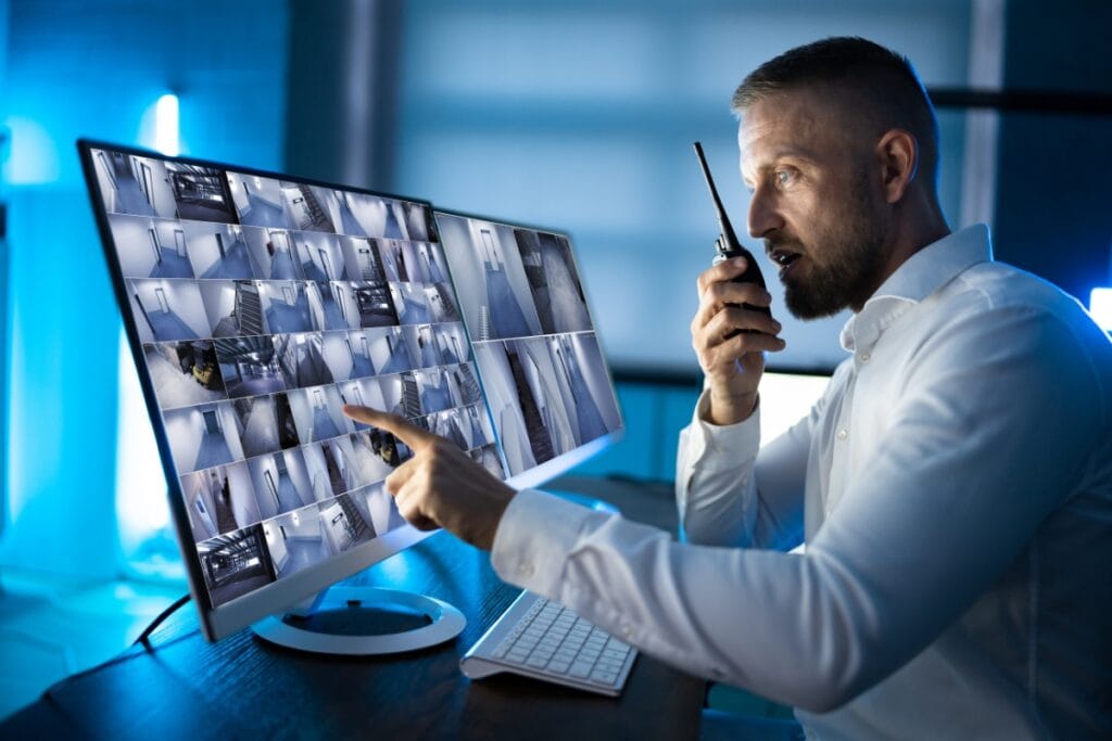 A man in a white shirt meticulously oversees multiple corporate security camera feeds on two large monitors while speaking into a walkie-talkie.