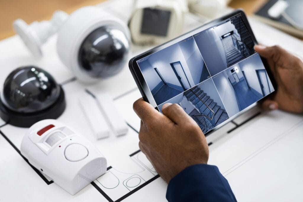 Hands holding a tablet displaying four security camera feeds over a table with CCTV cameras and a security alarm—integral elements of corporate security ensuring comprehensive protection.