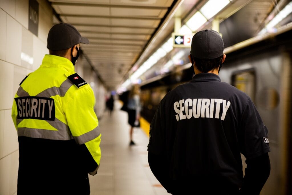 Two corporate security guards in uniform stand on a subway platform, facing the tracks as a train arrives.