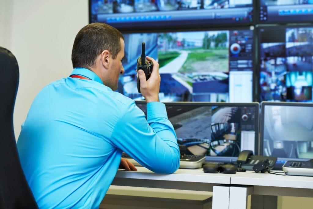 A man in a blue shirt sits at a desk, using a walkie-talkie, expertly managing corporate security as he monitors multiple surveillance screens displaying various camera feeds.