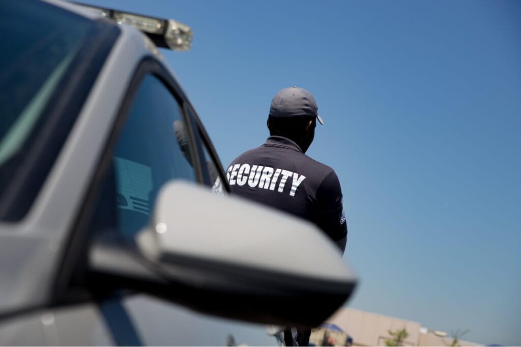 A corporate security guard stands near a vehicle, wearing a dark uniform with "Security" emblazoned on the back, under a clear blue sky.