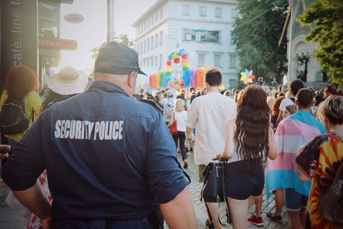 A crowd at a colorful street event bustles with excitement, while an unarmed security officer stands watchfully in the foreground.