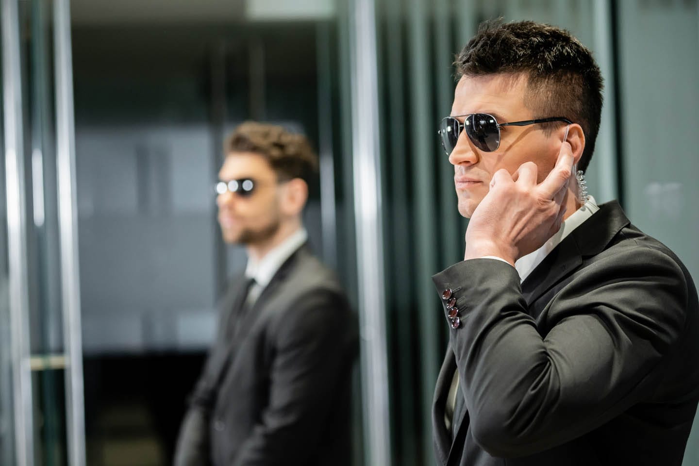 Two men in suits and sunglasses stand in an office setting, exuding an air of executive protection. The man in the foreground has an earpiece and is touching it, while the other stands watchfully in the background.