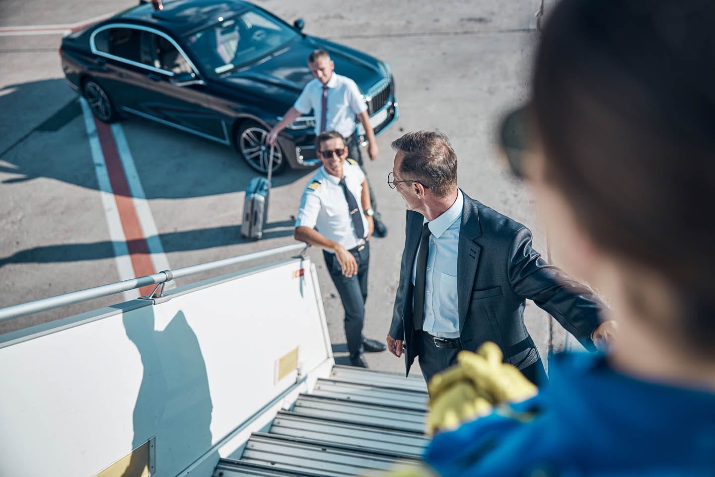 An executive in a suit climbs the airplane stairs, turning toward two uniformed pilots standing on the tarmac near a parked black car, ensuring every step aligns with his executive protection protocol.