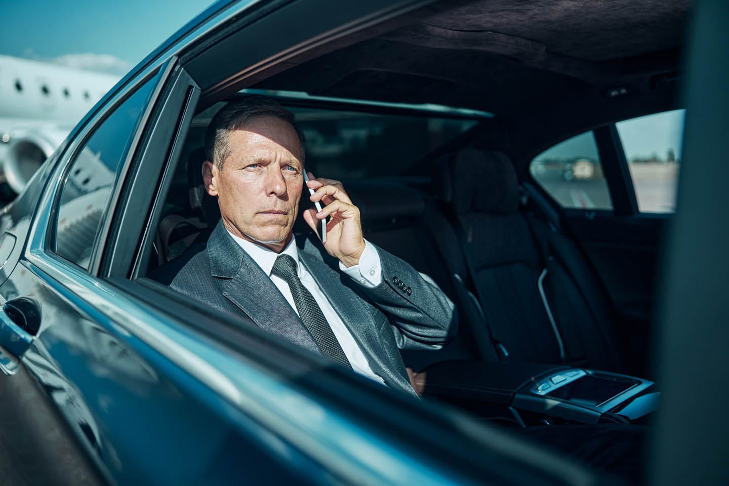 A man in a suit sits in a car, talking on a smartphone, with an airplane in the background, embodying executive protection and personal safety amidst his high-stakes environment.