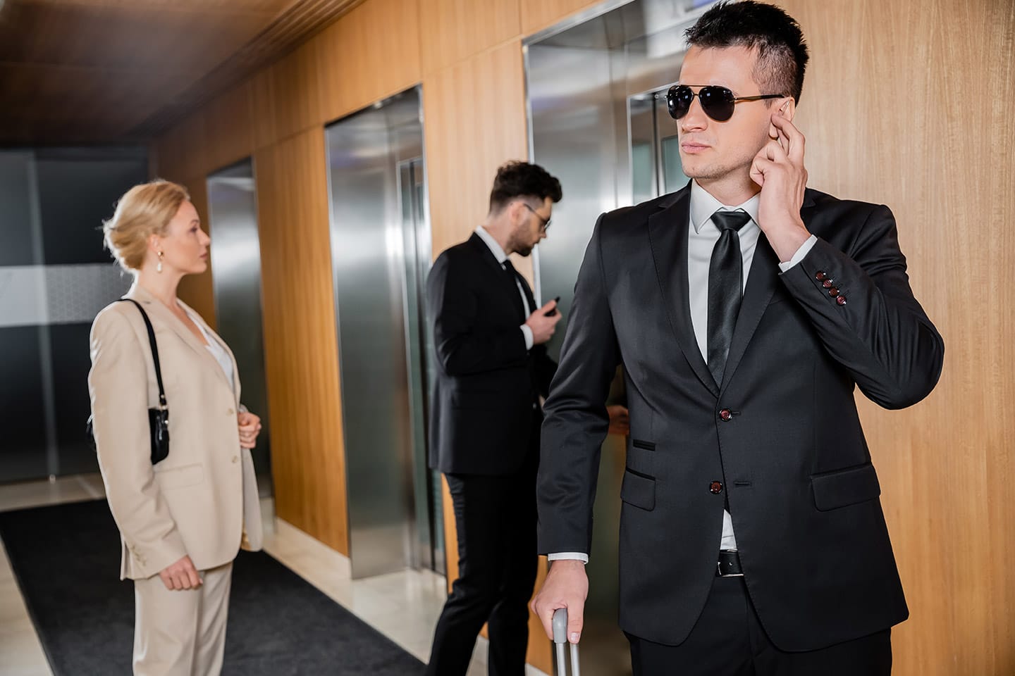 A man in a black suit and sunglasses, resembling unarmed security guards, listens intently. Another man checks his phone. A woman in a light suit observes. They stand near elevators in a wood-paneled hallway.