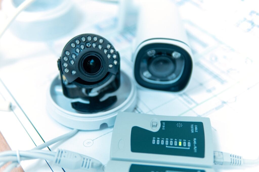 Close-up of security cameras, one disassembled with visible infrared LEDs, and a network cable tester on a technical blueprint, tools often used in family law investigations to ensure thorough and precise gathering of evidence.