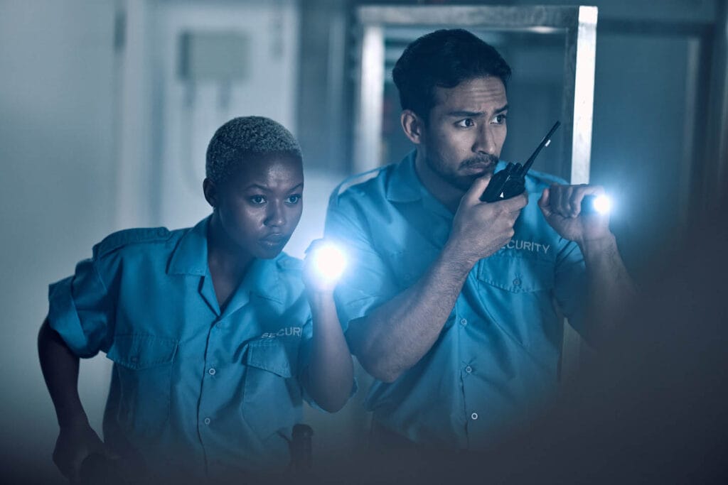 Two security guards in blue uniforms rove the dimly lit area, flashlights in hand and focused expressions. One guard communicates over a walkie-talkie as they diligently perform their patrol.