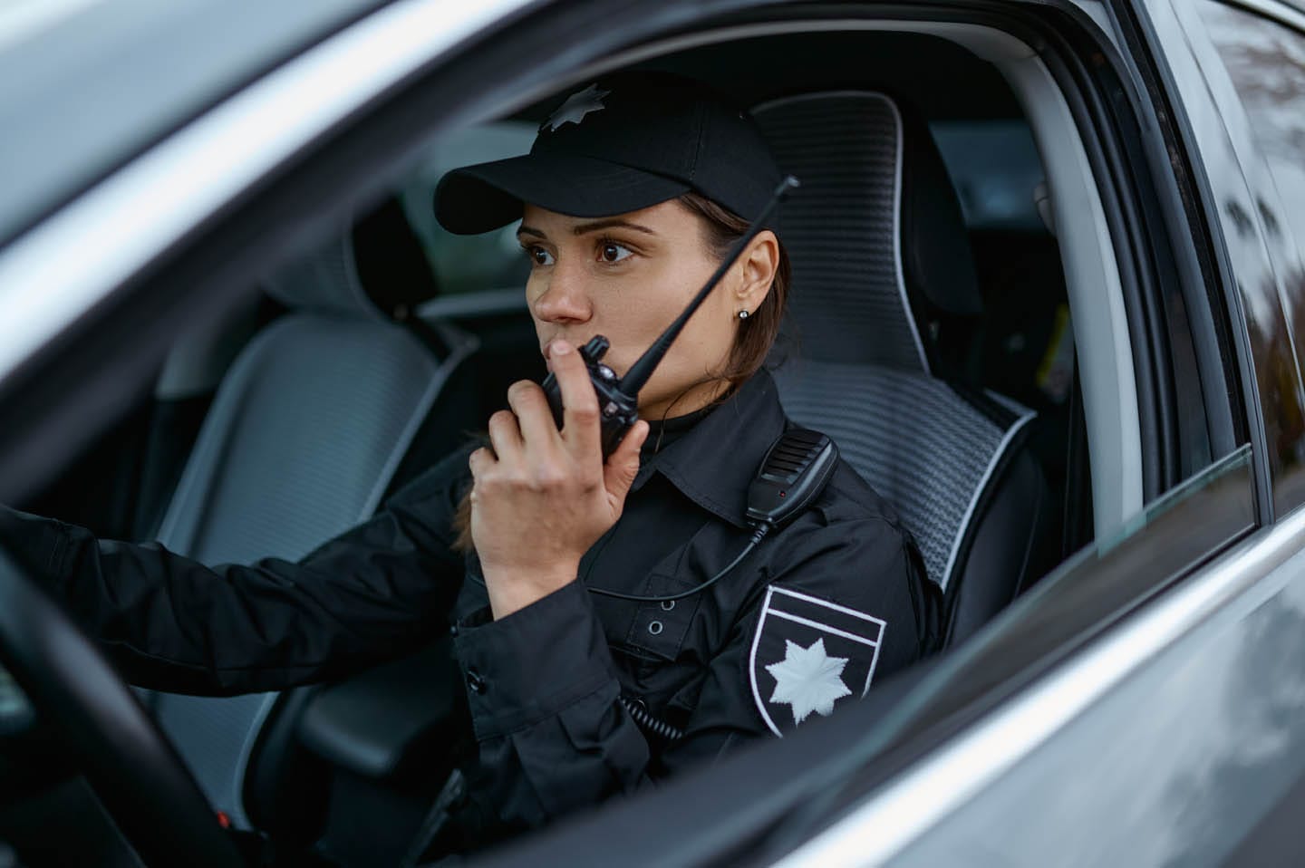 A police officer in uniform sits in a patrol car, speaking into a radio, conducting roving surveillance to enhance security in the area.