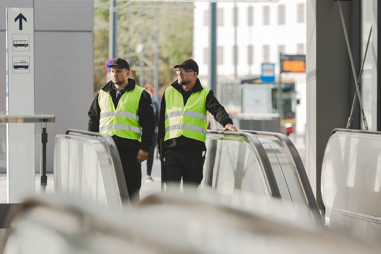 Two security personnel in reflective vests conduct mobile surveillance as they walk beside escalators in a modern transit station.