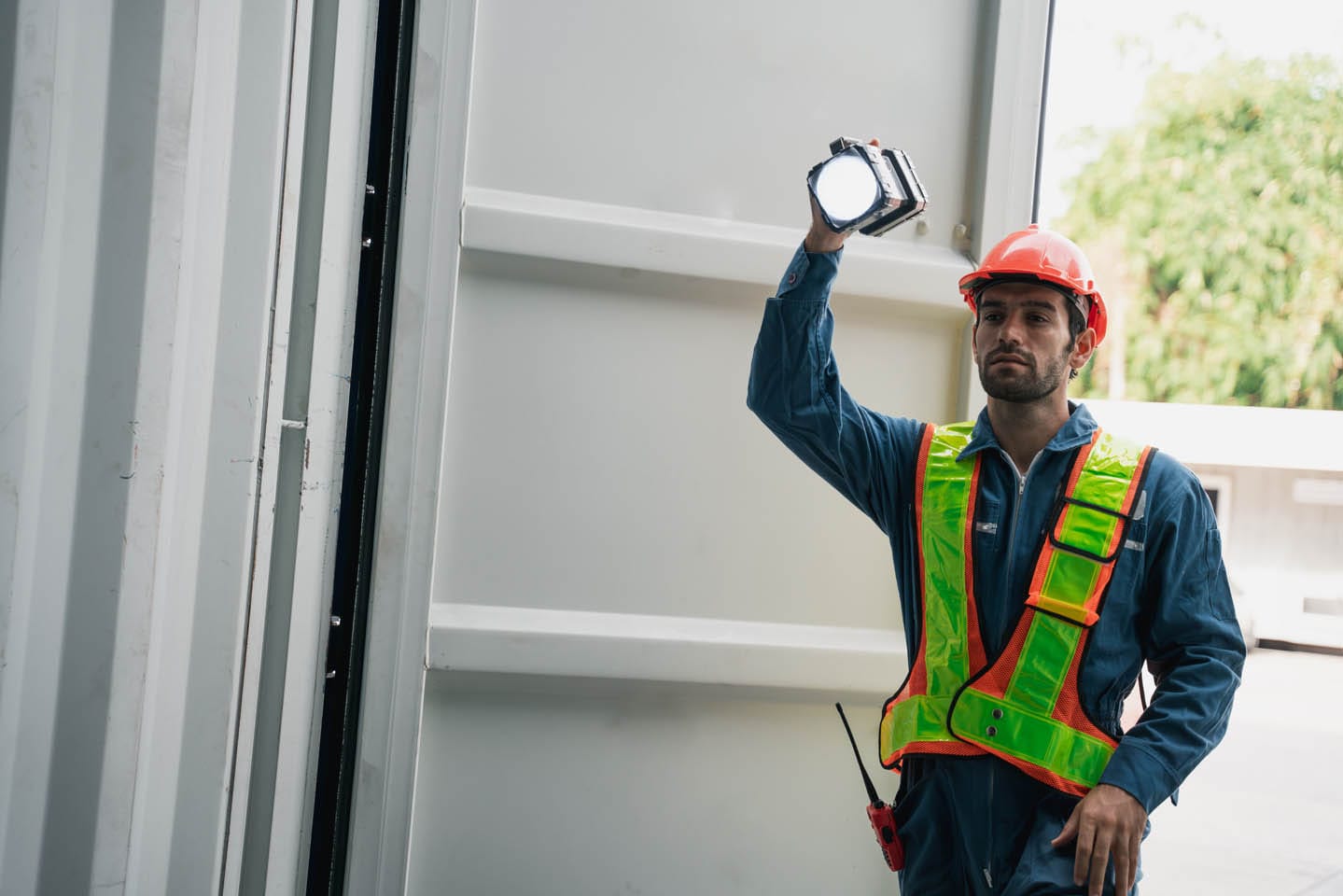 A worker in a hard hat and safety vest, part of the roving patrols, holds a flashlight, standing in an open doorway with trees visible outside.