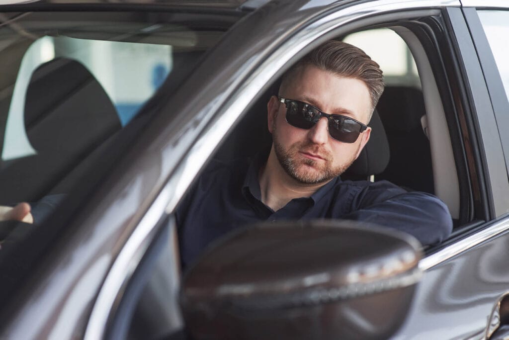 A man in sunglasses sits in the driver's seat of a car, looking out the window, as if on roving patrols through familiar streets.