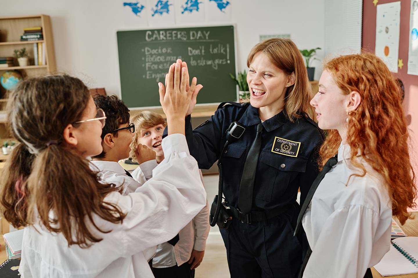 During Career Day, a police officer high-fives a student in the classroom, discussing vital topics like Active Shooter Response training as other students watch eagerly. In the background, a chalkboard lists various career paths to inspire curiosity and ambition.