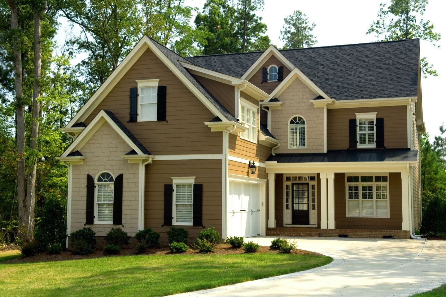 A two-story suburban house with brown and beige siding, black shutters, a dark roof, and a driveway. Surrounded by trees and greenery, it has a covered front porch and multiple windows—perfect for active threat training scenarios.